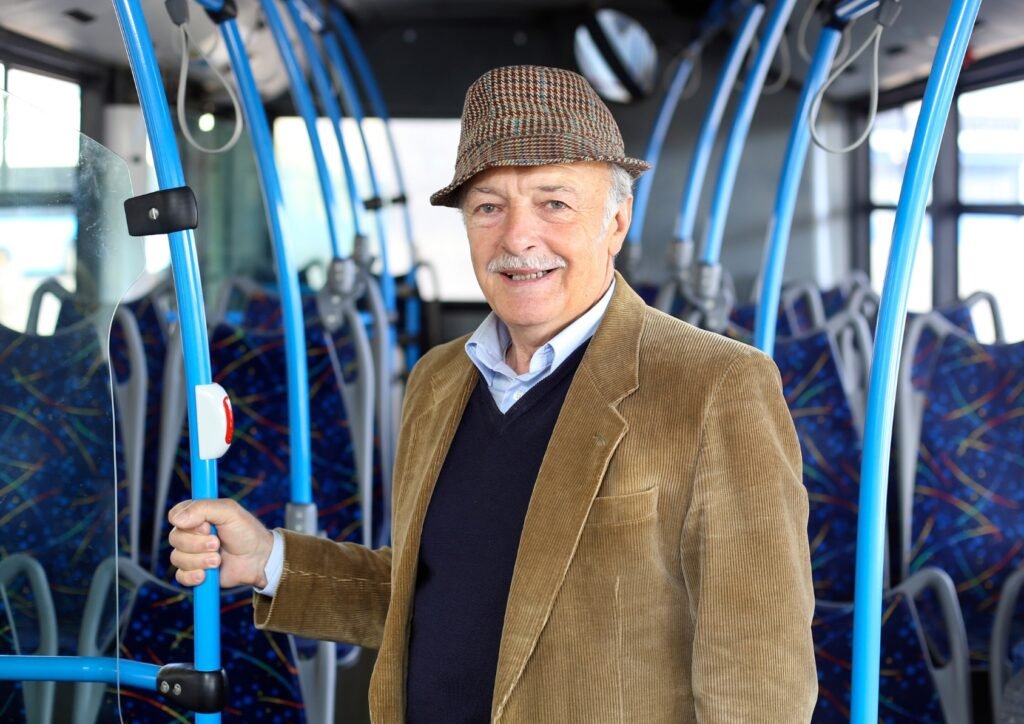 Smiling senior man in tweed jacket and cap riding public bus, holding handrail while standing in the aisle